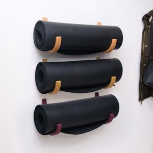 Op de afbeelding: Drie opgerolde zwarte yogamatten zijn aan een witte muur bevestigd. Elke mat wordt vastgehouden door een houten houder met een gebogen ontwerp. De bovenste twee houders zijn lichtbruin en de onderste is dieppaars.