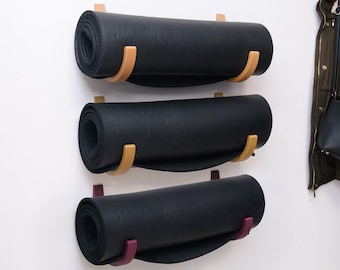 May include: Three rolled-up black yoga mats are mounted on a white wall. Each mat is held by a wooden holder with a curved design. The top two holders are light brown, and the bottom one is a deep purple.