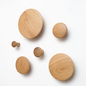 Può includere: Un insieme di ganci da parete in legno di varie dimensioni. I ganci sono realizzati in legno chiaro e presentano un motivo a venature naturali. Il design include forme circolari e cilindriche, creando un'estetica moderna e minimalista.