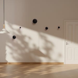 Op de afbeelding: Een minimalistische interieurscène met een reeks zwarte, bolvormige wandhaken van verschillende afmetingen, gemonteerd op een neutraal gekleurde muur. De haken zijn in een verspringend patroon gerangschikt. Een witte deur en een hanglamp zijn ook zichtbaar.