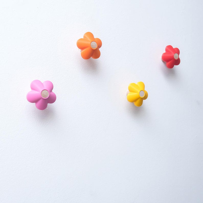 May include: Four wall hooks shaped like five-petaled flowers in bright colors. The set includes one large pink hook, one orange hook, one yellow hook, and one smaller red hook. Each flower-shaped hook features a circular center detail.