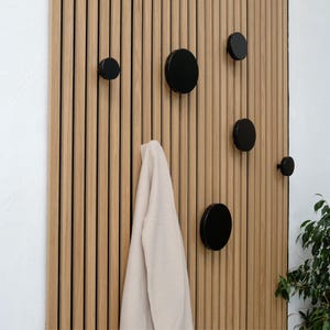 Könnte beinhalten: Eine Holzwand mit vertikalen Latten, die mit schwarzen runden Haken ausgestattet ist. Ein beiger Pullover hängt an einem der Haken.