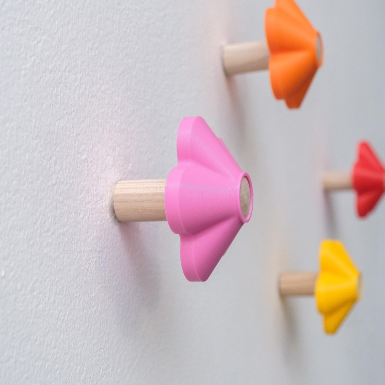 May include: A set of wall-mounted hooks featuring a cylindrical wooden base and a star-shaped front piece in vibrant colors including pink, orange, yellow, and red, attached to a white surface.