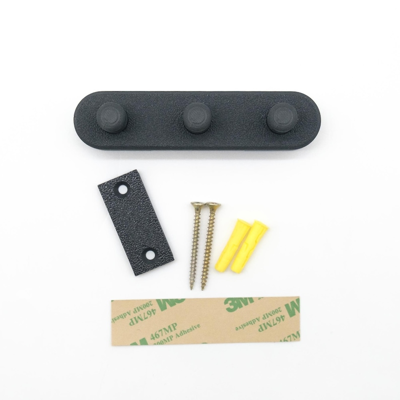 May include: A black wall hook with three knobs, a small mounting plate, screws, wall plugs, and adhesive tape. The hook is designed for hanging items and is made of a durable material. The hardware is included for easy installation.