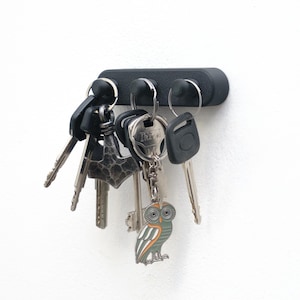 May include: A black magnetic key holder with various keys hanging from it. The keys include a decorative owl keyring. The holder is mounted on a white wall. The key holder is approximately 10 cm in length.