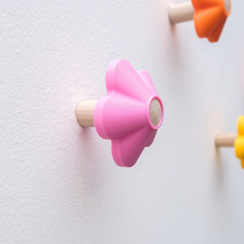 May include: A wall-mounted hook shaped like a five-petaled flower with a bright pink finish and a natural light wood cylindrical base. The hook extends outward from the wall, featuring a smooth, matte surface and a rounded center detail.