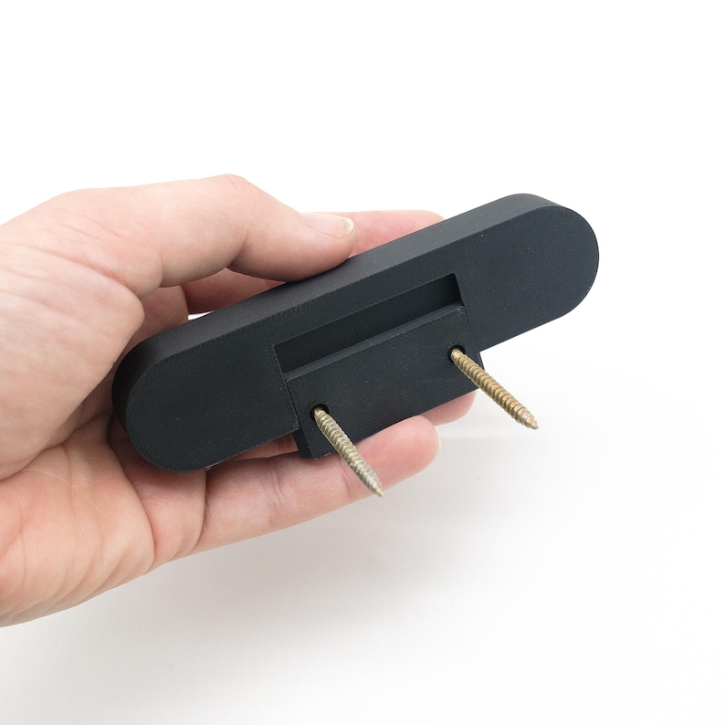 May include: A black, rectangular wooden object with rounded ends, featuring a recessed area and two gold-coloured screws. The object is held in a hand against a white background.