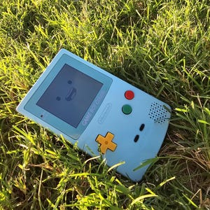 Puede incluir: Una consola de juegos portátil Game Boy Color azul claro con una cara sonriente en la pantalla, colocada en hierba verde. La consola tiene botones rojos y verdes, una cruceta amarilla y el logotipo de Nintendo.