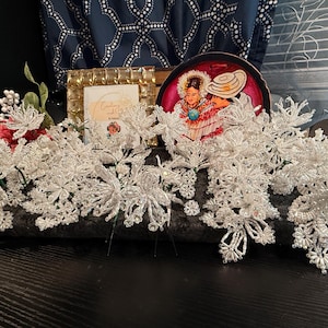 May include: A display of handmade, white beaded hair accessories, including floral and snowflake designs. A framed picture and a decorative plate with a couple in traditional attire are also visible. The items are arranged on a dark surface.