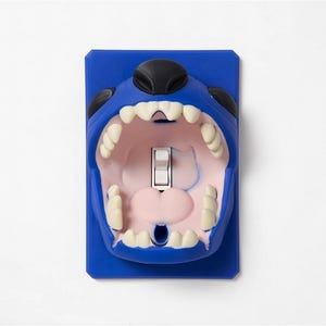 May include: A novelty light switch cover in the shape of a cartoon character's open mouth. The cover is blue with white teeth, a pink interior, and a black nose. The switch is white.