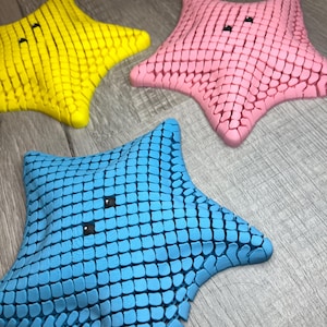 May include: Three colorful starfish toys in yellow, pink, and blue. Each starfish has a textured surface and two small black eyes. The toys are arranged on a wooden surface.