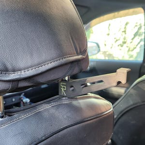 May include: A black car seat headrest with a black metal hook attached to it. The hook is labeled "CR".