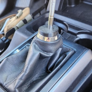 May include: A close-up of a black leather gear shift knob with a silver metal top and a threaded rod. The knob is installed in a car's center console.