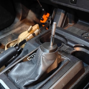 May include: A close-up of a car's gear shift assembly. The shift lever is chrome and black, with a black leather boot. The assembly is set in a black plastic console. The background shows other car interior details.
