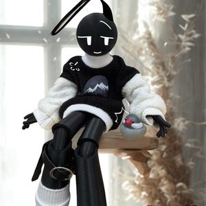 Puede incluir: Muñeca en blanco y negro con cabeza redonda, pequeños cuernos y cara sonriente. La muñeca lleva un suéter negro con un gráfico de montaña y mangas blancas, pantalones negros y botas negras. Una pequeña figura de pájaro está sobre una estantería de madera.