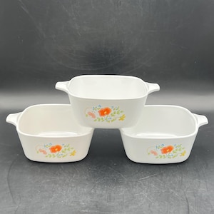 May include: Three white square casserole dishes with handles. Each dish features a floral design with orange and yellow flowers and green leaves. The dishes are stacked on a dark surface.