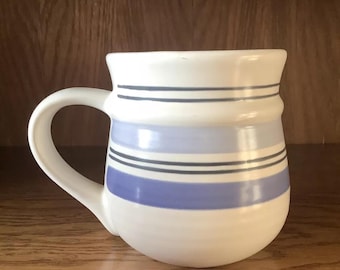 May include: A white ceramic mug with a blue and gray striped design. The mug has a rounded body and a handle.