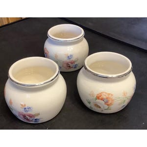 May include: Three white ceramic jars with floral designs. The jars have a silver rim around the top and feature a variety of flowers in shades of pink, blue, and orange.