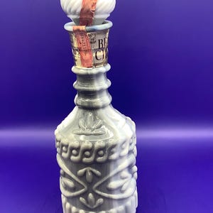 May include: A white and gray glass decanter with a swirl design and a white stopper. The decanter has a label that reads "BEACH CINNAMON" and "211246".