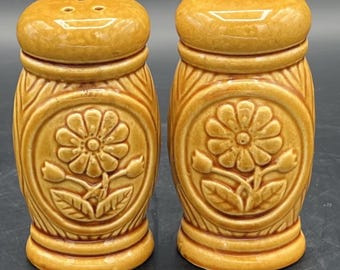 May include: A pair of brown ceramic salt and pepper shakers with a floral design. The shakers have a raised design of a flower with leaves and a textured pattern on the sides.