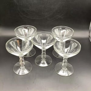 May include: Set of five clear glass champagne coupes with beaded stems. The coupes are arranged in a semi-circle on a dark surface.