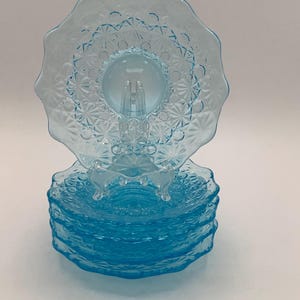 May include: A set of five light blue pressed glass plates featuring a scalloped edge and a repeating starburst pattern. One plate is displayed upright on a clear plastic stand, while the remaining four plates are stacked neatly beneath it.