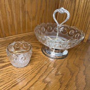 May include: Two-piece glass serving set. The larger piece is a tiered serving dish with a silver handle and base. The smaller piece is a matching bowl. Both pieces have a scalloped edge design and gold trim.