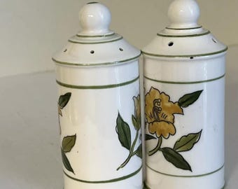 May include: A pair of white ceramic salt and pepper shakers with green and yellow floral designs. The shakers have a round top and a cylindrical body.
