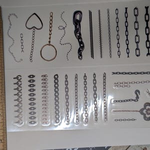 May include: Clear sheet of chain-themed stickers. Designs include various chain styles, a heart-shaped charm, and a flower-shaped charm. The stickers are in shades of silver, black, and gold. A wooden ruler is in the image.