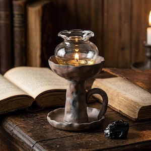 May include: A clear glass oil lamp with a lit wick sits atop a rustic, brown metal stand. The lamp has a round, ruffled top and a small handle. The background features open books and a candle.
