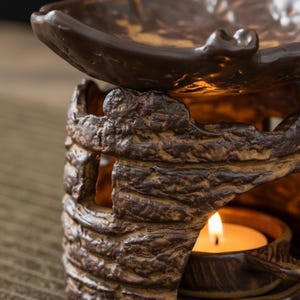 May include: A brown ceramic essential oil diffuser with a textured, bark-like design. The top bowl holds oil, and a lit tea light candle inside provides heat. The warm glow of the candle illuminates the intricate details of the diffuser.