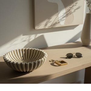 May include: A beige, ribbed ceramic bowl sits on a light wood surface, next to sunglasses and a key chain. The bowl has a scalloped edge and a textured interior. A vase and artwork are in the background.