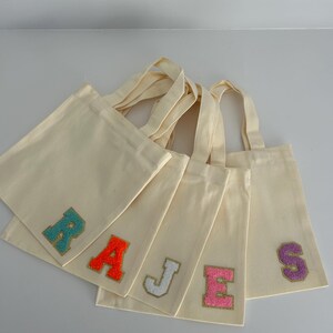 May include: Cream canvas tote bags with embroidered letters spelling "RAJS". The letters are teal, orange, white, pink, and purple, with gold trim. Each bag has a cream handle. Ideal for carrying items.