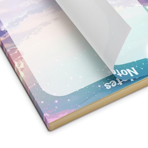May include: A notepad with a dreamy sky design, featuring pastel pink, purple, and blue hues. The notepad has a gold-colored edge and the word "Notes" is visible on the side. A page is being peeled back, revealing a blank white surface.