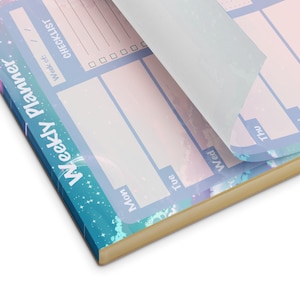 May include: A blue and pink weekly planner with a galaxy design. The planner has a checklist, a section for each day of the week, and a space for notes. The planner is open to the Monday page.