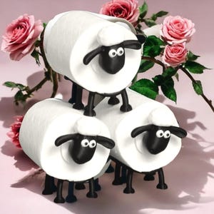 May include: Three novelty sheep ornaments with white bodies and black faces, ears, and legs are stacked. Pink roses and green leaves are arranged behind the sheep against a pink backdrop. The sheep have a cartoonish design.