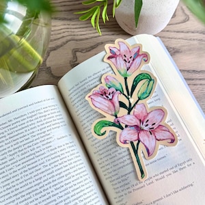 May include: A die-cut bookmark featuring a hand-drawn illustration of three pink lily flowers with green stems and leaves. The bookmark is placed inside an open book with visible printed text on the pages, resting on a light wooden surface.