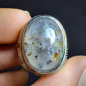 Pode incluir: Um anel de prata com uma pedra grande de forma oval. A pedra é uma ágata cinza claro com inclusões marrons e pretas. O anel tem uma banda texturizada.
