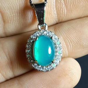 Puede incluir: Un collar con colgante de plata con una piedra preciosa ovalada de color turquesa. La piedra preciosa est&aacute; rodeada por un halo de peque&ntilde;os cristales transparentes. El colgante cuelga de una fina cadena de plata.