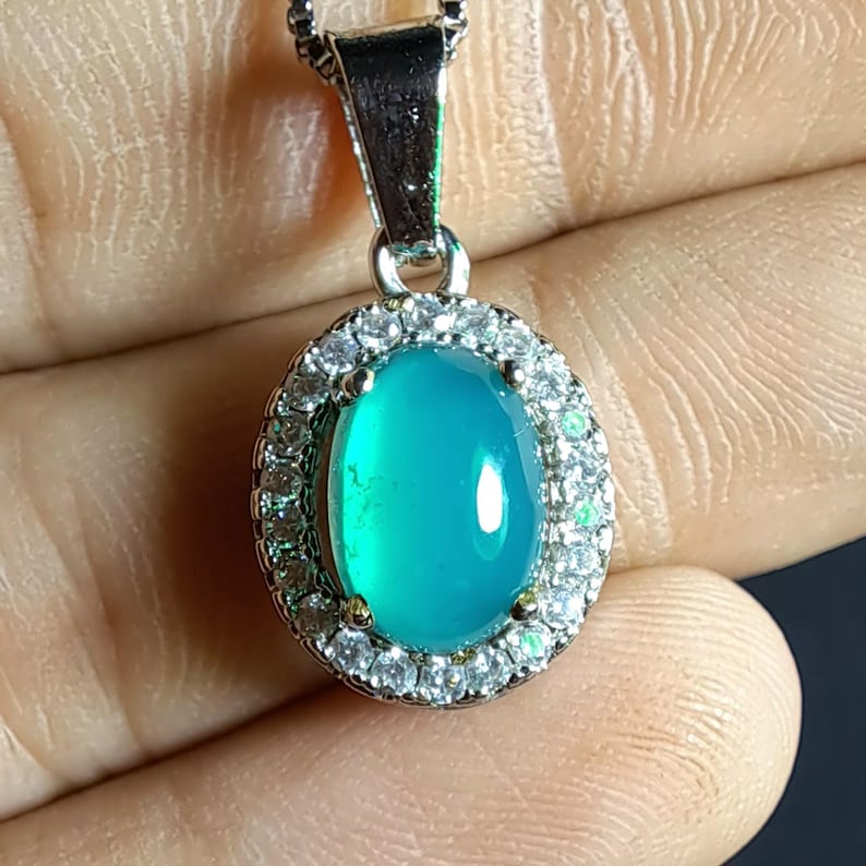 Puede incluir: Un collar con colgante de plata con una piedra preciosa ovalada de color turquesa. La piedra preciosa est&aacute; rodeada por un halo de peque&ntilde;os cristales transparentes. El colgante cuelga de una fina cadena de plata.