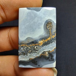 Puede incluir: Una gema rectangular con un patrón similar a un paisaje. La piedra presenta capas de tonos grises, blancos, negros y dorados, creando un diseño natural único. La superficie pulida muestra los intrincados detalles de la formación de la piedra.