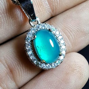 Puede incluir: Un collar con colgante de plata con una piedra preciosa ovalada de color turquesa. La piedra preciosa est&aacute; rodeada por un halo de peque&ntilde;os cristales transparentes. El colgante se muestra sobre un fondo neutro.