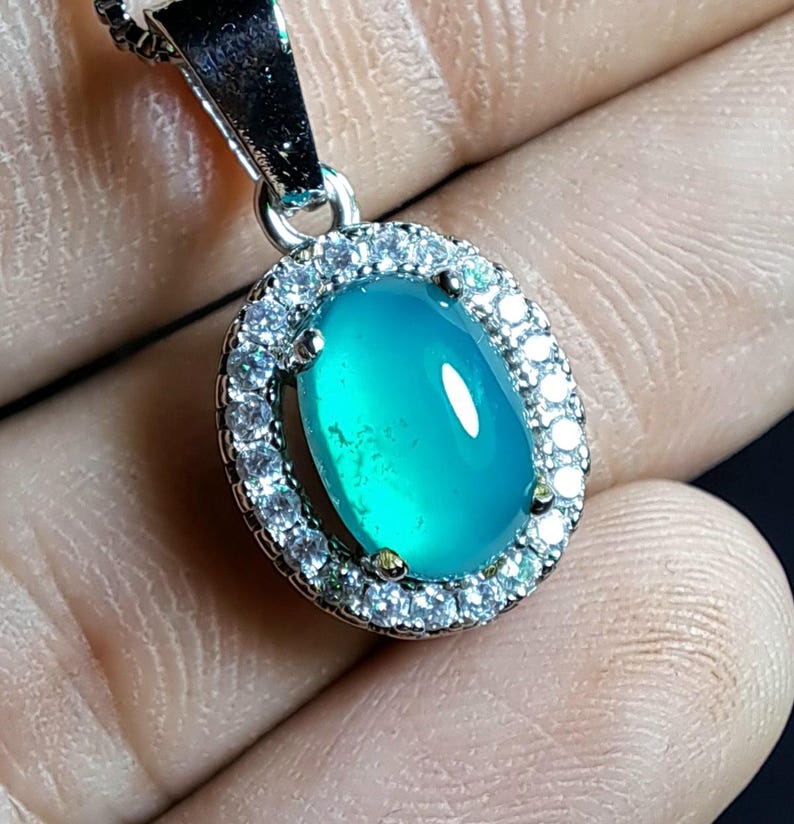 Puede incluir: Un collar con colgante de plata con una piedra preciosa ovalada de color turquesa. La piedra preciosa est&aacute; rodeada por un halo de peque&ntilde;os cristales transparentes. El colgante se muestra sobre un fondo neutro.