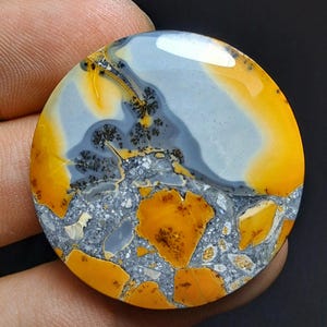 May include: A round, polished gemstone with a unique pattern. The stone features a blend of colors, including gray, yellow, and orange, with intricate black dendritic inclusions. The overall design is a mix of smooth and textured areas.