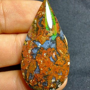 Può includere: Un cabochon di agata lucido a forma di lacrima con una miscela di colori marrone, arancio e bianco. La pietra presenta un motivo unico e vorticoso e un bagliore iridescente verde.