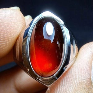 Puede incluir: Un anillo de plata con una gema grande, ovalada y de color rojo intenso. La gema pulida refleja la luz. La banda del anillo es de plata pulida, con un diseño sencillo. El diseño del anillo es clásico y elegante.