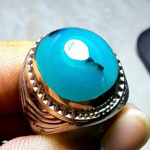 Pode incluir: Um anel de prata com uma pedra turquesa. O anel tem um design detalhado com uma banda texturizada e uma borda decorativa em torno da pedra.