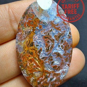 May include: An oval gemstone displaying a rich tapestry of colours. The stone's surface is a blend of orange, brown, blue, and white, forming a distinctive, textured design. The text "US IMPORT TARIFF FREE GUARANTEE" is stamped on the top right corner.