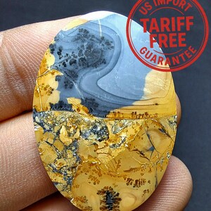 Może przedstawiać: Owalny kamień szlachetny o unikalnym wzorze. Górna połowa ma jasnoniebieski i szary wzór wiru, a dolna połowa prezentuje mieszankę żółtych, brązowych i czarnych tekstur. Kamień ma naturalny, ziemisty wygląd. Widoczny jest tekst "US Import Tariff Free Guarantee".