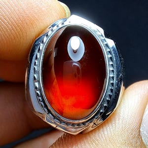 May include: A silver ring featuring an oval, deep red gemstone. The polished gemstone reflects light, displaying a colour gradient. The ring's setting has a detailed, textured design around the stone.